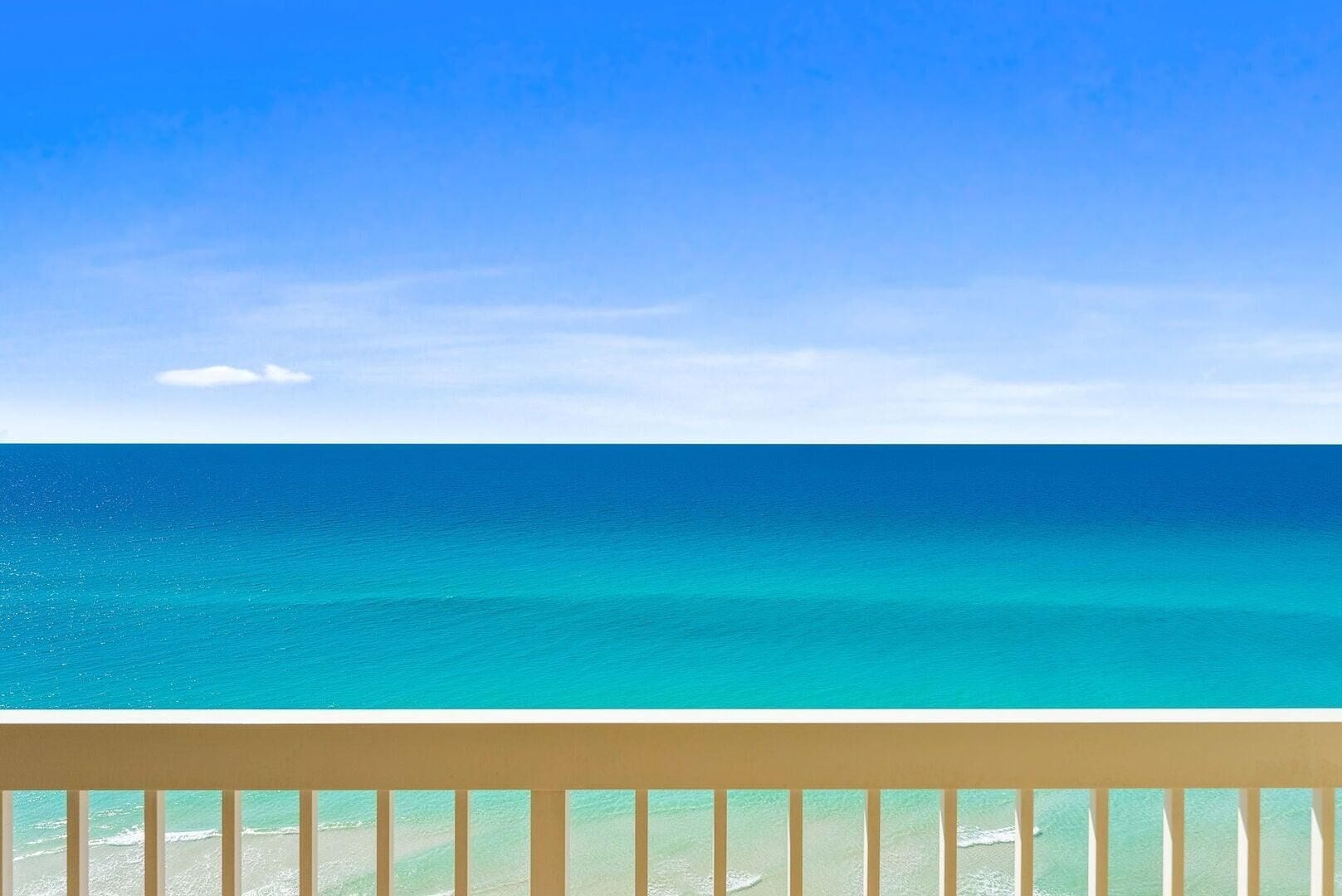 A clear view of a turquoise ocean from a balcony with a wooden railing.
