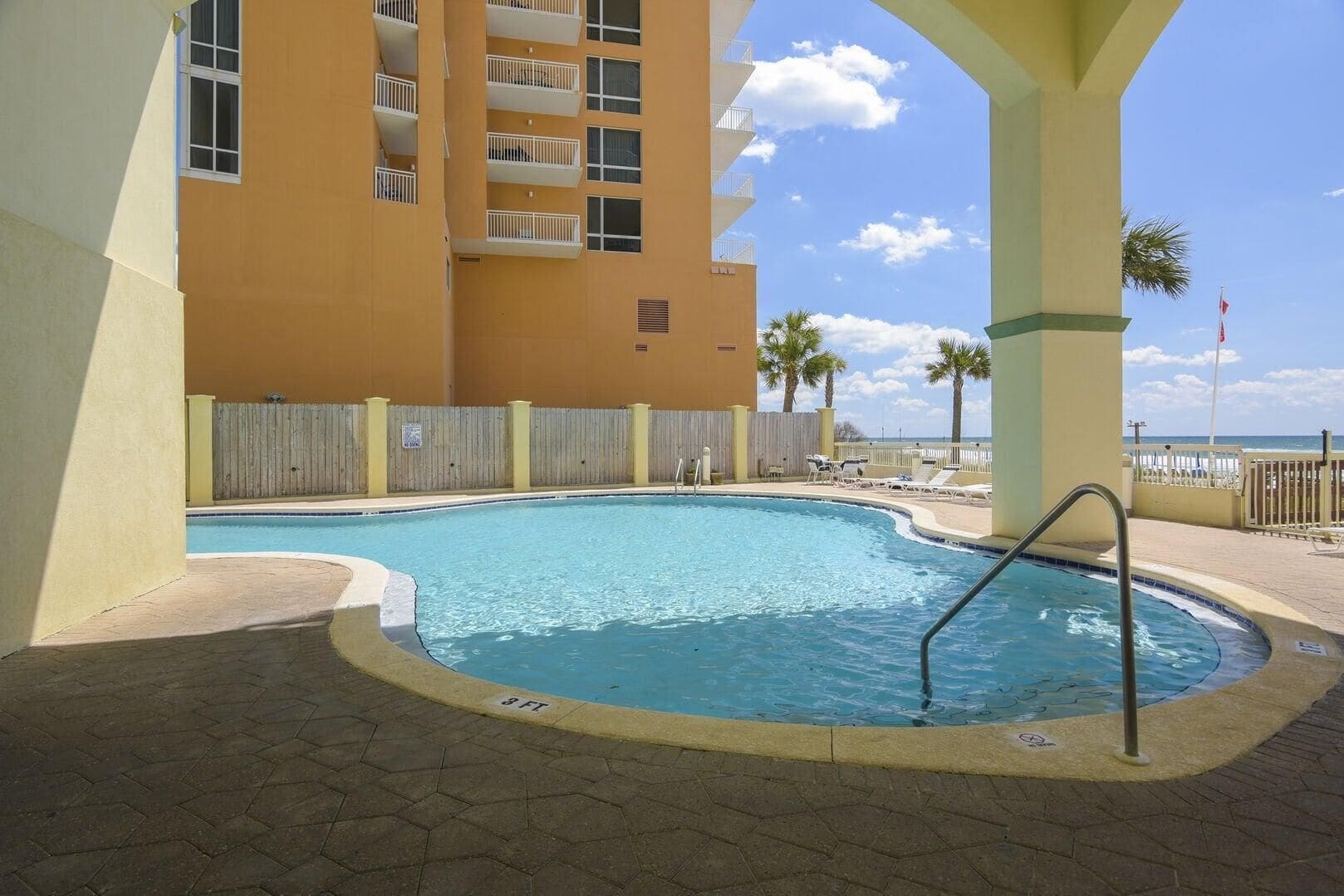 Une piscine avec un jacuzzi encastré, entourée d’une clôture en bois, et un bâtiment visible au loin.