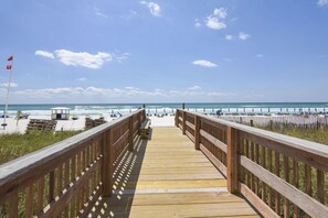 Ein hölzerner Steg führt zu einem Strand mit Sonnenschirmen und Liegestühlen.