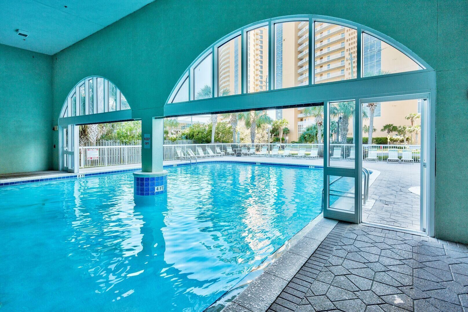 Une piscine intérieure avec des parois en verre transparent, un sol carrelé et une vue sur un quartier résidentiel.