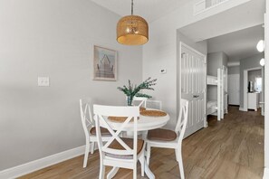 Un coin repas avec une table et des chaises blanches, un luminaire suspendu en fibres tressées et un tableau encadré au mur.