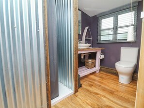 A bathroom with a wooden floor, a white toilet, a sink with a mirror, and a window with blinds. - Hedgehogs Lodge (South Molton)