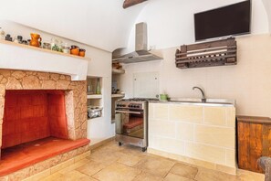 A compact kitchen with a stone fireplace, a built-in oven, a stove, and a wooden shelf above the counter. - Trullo Sardella by BarbarHouse (Ceglie Messapica)