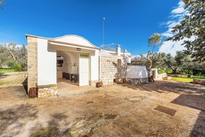 A white building with a stone facade, a tiled roof, and a small garden area with trees and a paved path. - Trullo Sardella by BarbarHouse (Ceglie Messapica)