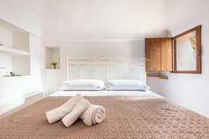 Una camera da letto con un letto, cuscini, un asciugamano arrotolato, un armadietto in legno e una mensola a muro.