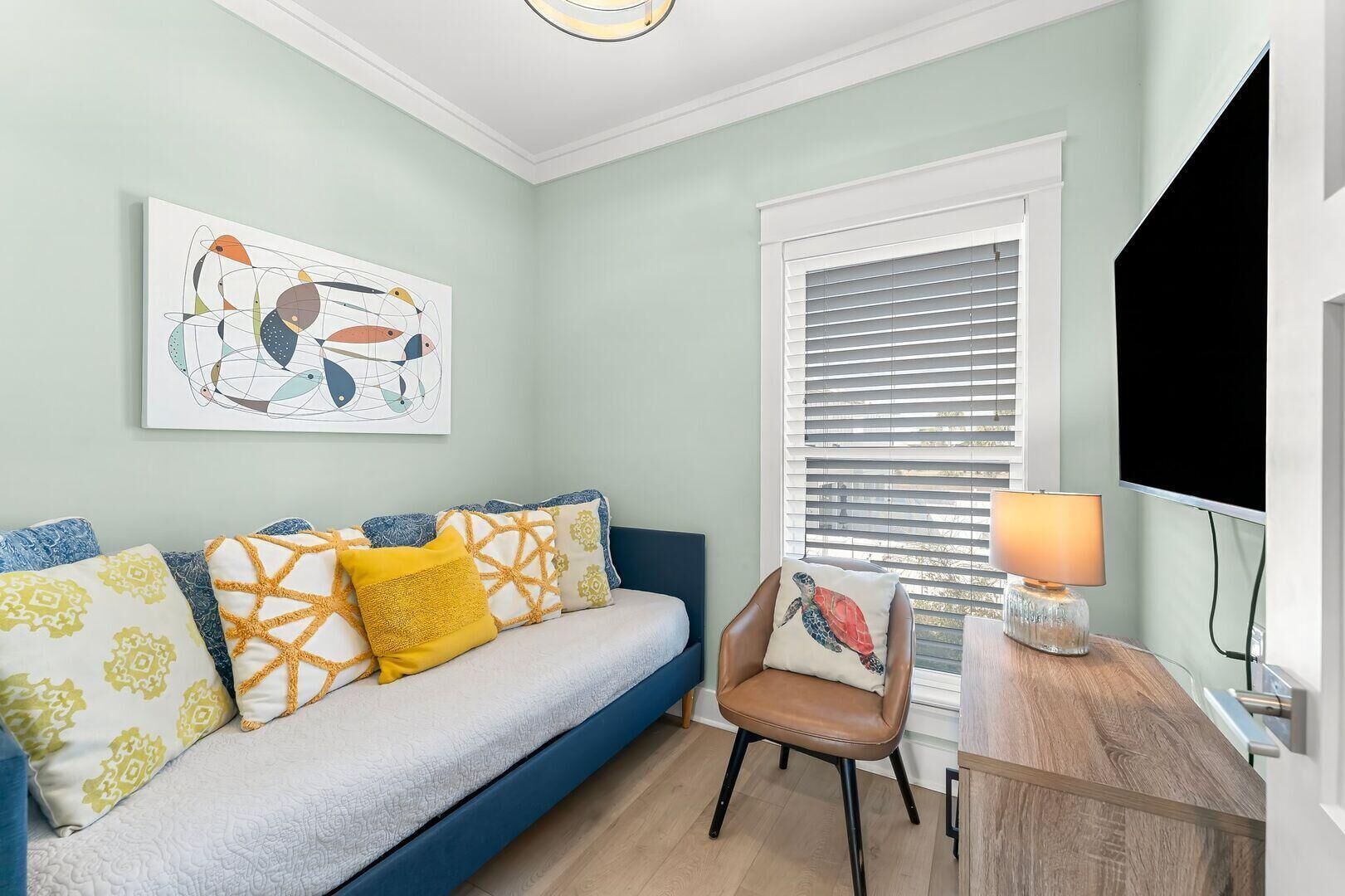 Habitación con sofá azul, cojines estampados, almohada con motivos de tortuga, mesita de madera con lámpara y televisor grande.
