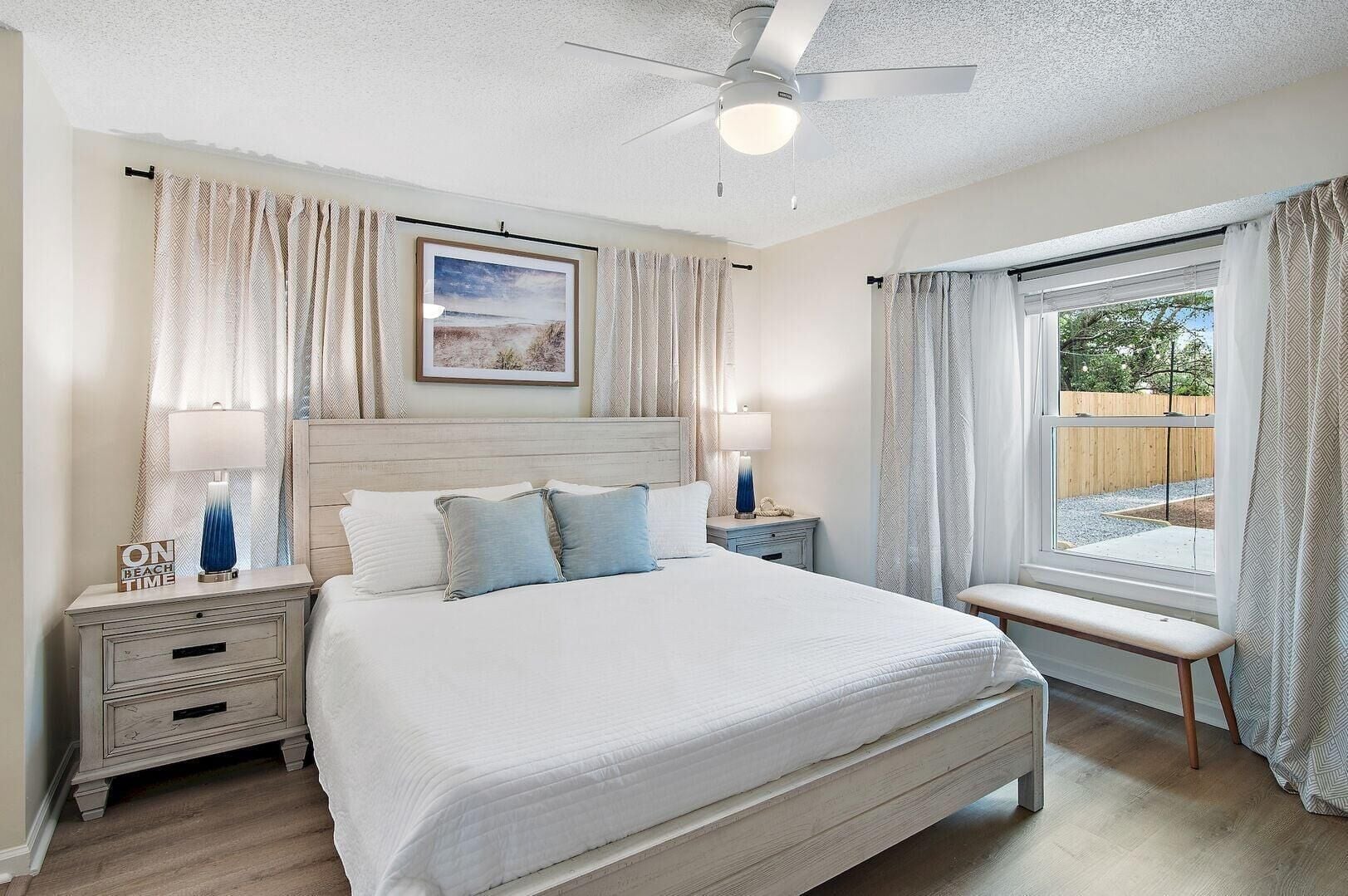 A bedroom with a bed, bedside tables, a ceiling fan, a window with curtains, and a framed picture on the wall.