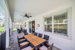 Um pátio externo com mesa e cadeiras de madeira, um ventilador de teto e amplas janelas com persianas.