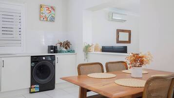 A compact kitchen with a washing machine, a dining table with chairs, and a small living area with a TV and a framed picture on the wall.