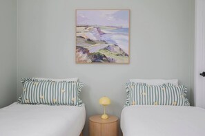 Ein Zimmer mit zwei Betten, jedes mit gestreiften Kissen, einem runden Nachttisch mit Lampe und einem Landschaftsbild an der Wand.