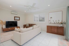 Ein Wohnzimmer mit Fernseher, braunem Ledersofa, weißem Sofa, Holzschrank und Deckenventilator.