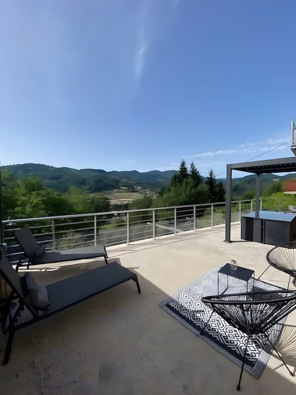 A balcony with a view of hills, a seating area with a patterned rug, and a metal chair.