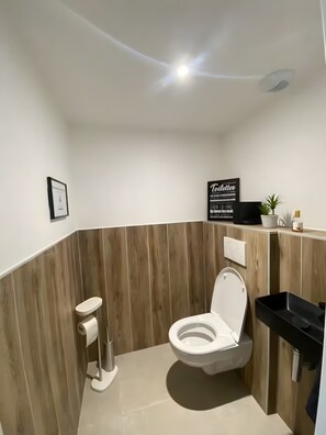 A modern bathroom with a corner toilet, a sink, a framed picture, and a sign on the wall.