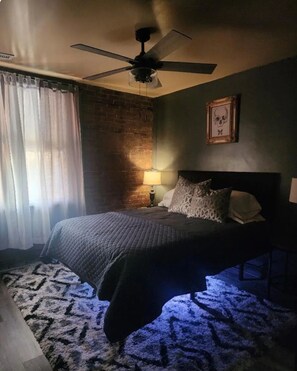 Um quarto com uma cama, um ventilador de teto, uma luminária e uma parede de tijolos.