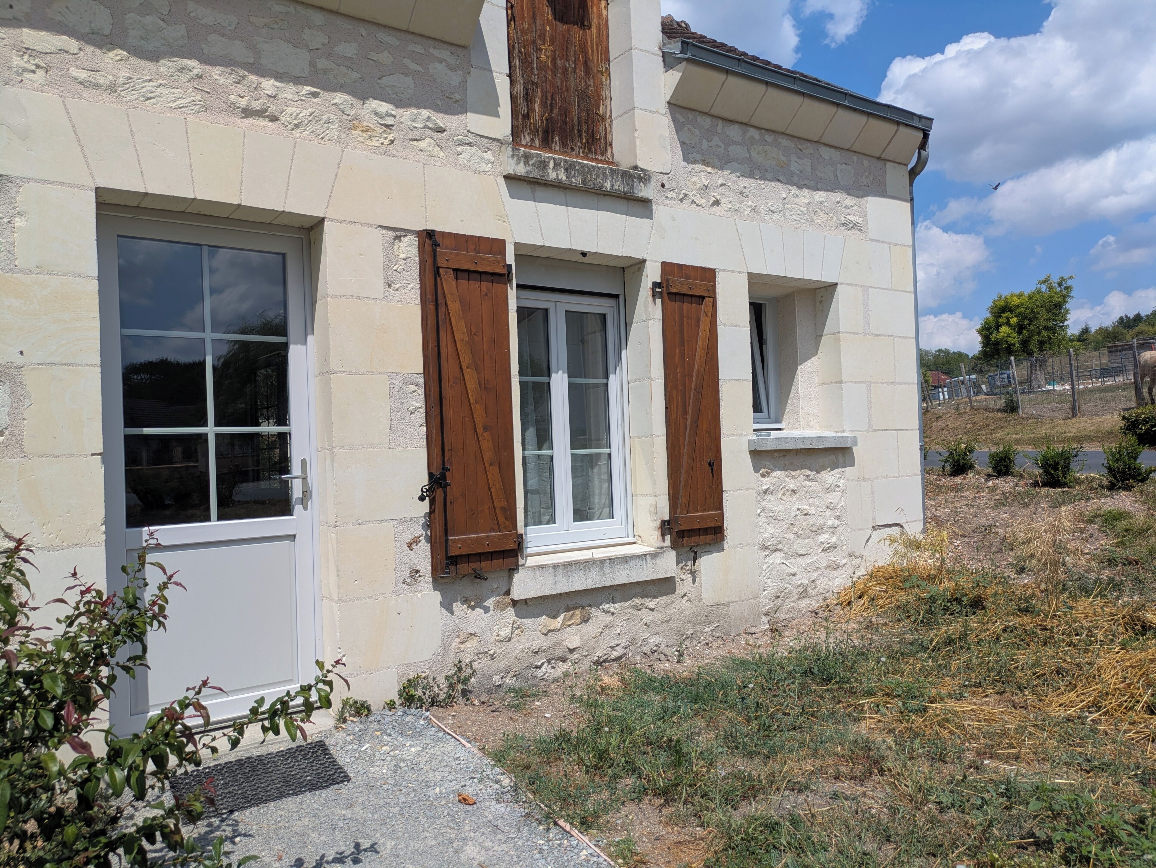 A stone building with a window and shutter, surrounded by dry grass and a fence.