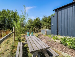 Ein Holztisch für Picknicks mit zwei Laternen, umgeben von einem Garten mit Rindenmulch und Pflanzen, und ein Metallschuppen im Hintergrund.