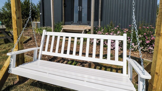 Un banc-balançoire en bois peint en blanc, doté de chaînes en métal, est placé devant un bâtiment à la façade sombre et au porche en bois.