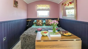 Une pièce chaleureuse avec un lit, une fenêtre ornée de rideaux à motifs floraux, un miroir décoré de motifs en forme de cœur et une table sur laquelle se trouvent un bol, un magazine et des pâtisseries.
