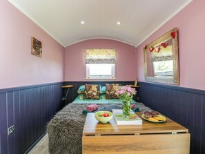 Ein gemütliches Zimmer mit Bett, einem Fenster mit floralem Vorhang, einem Spiegel mit Herzdekorationen und einem Tisch mit Schüssel, Zeitschrift und Gebäck.
