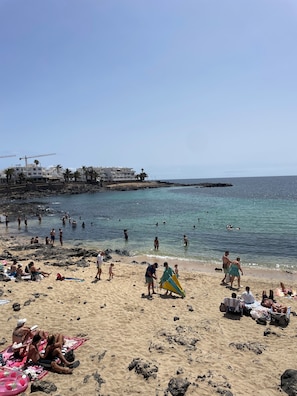 A beach with people sunbathing and swimming, a rocky shoreline, and buildings in the background. - BEAUTIFUL DUPLEX WITH A TERRACE IN COSTA TEGUISE (Costa Teguise)