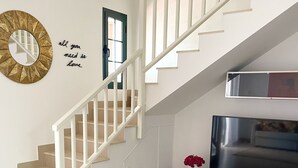 Una escalera con barandillas blancas que lleva a una plataforma, un televisor de pantalla plana sobre un soporte oscuro y un espejo decorativo en la pared.