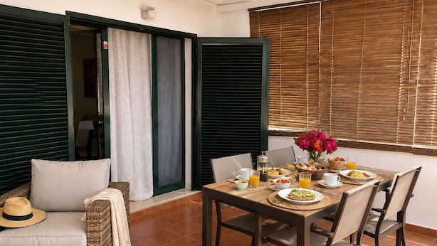 Un comedor con una mesa lista para el desayuno, una silla de mimbre y vistas a través de una ventana con persianas.
