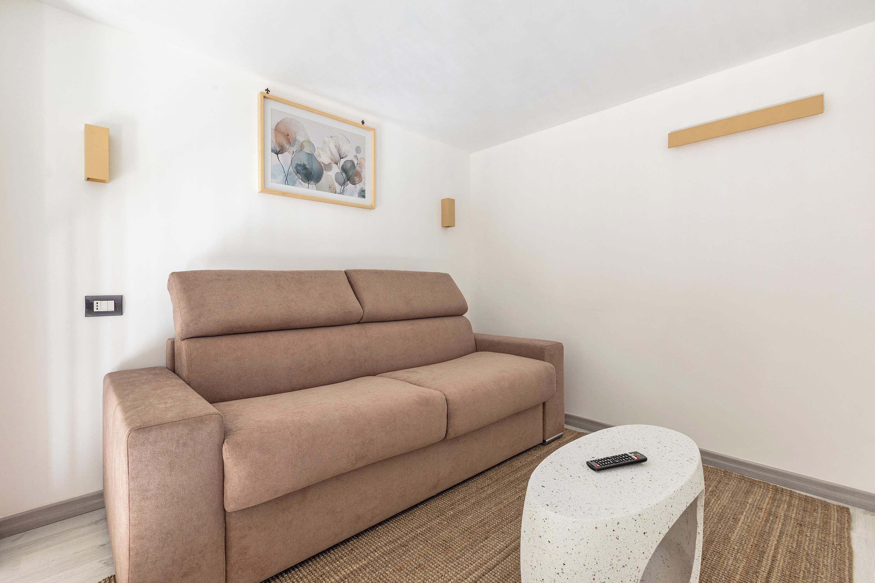 Uma sala de estar moderna com um sofá marrom, uma mesa redonda pequena com um controle remoto e um quadro na parede.