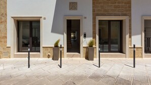 A building entrance with three doors, each with a number above it. There are potted plants near the doors and bollards in front.