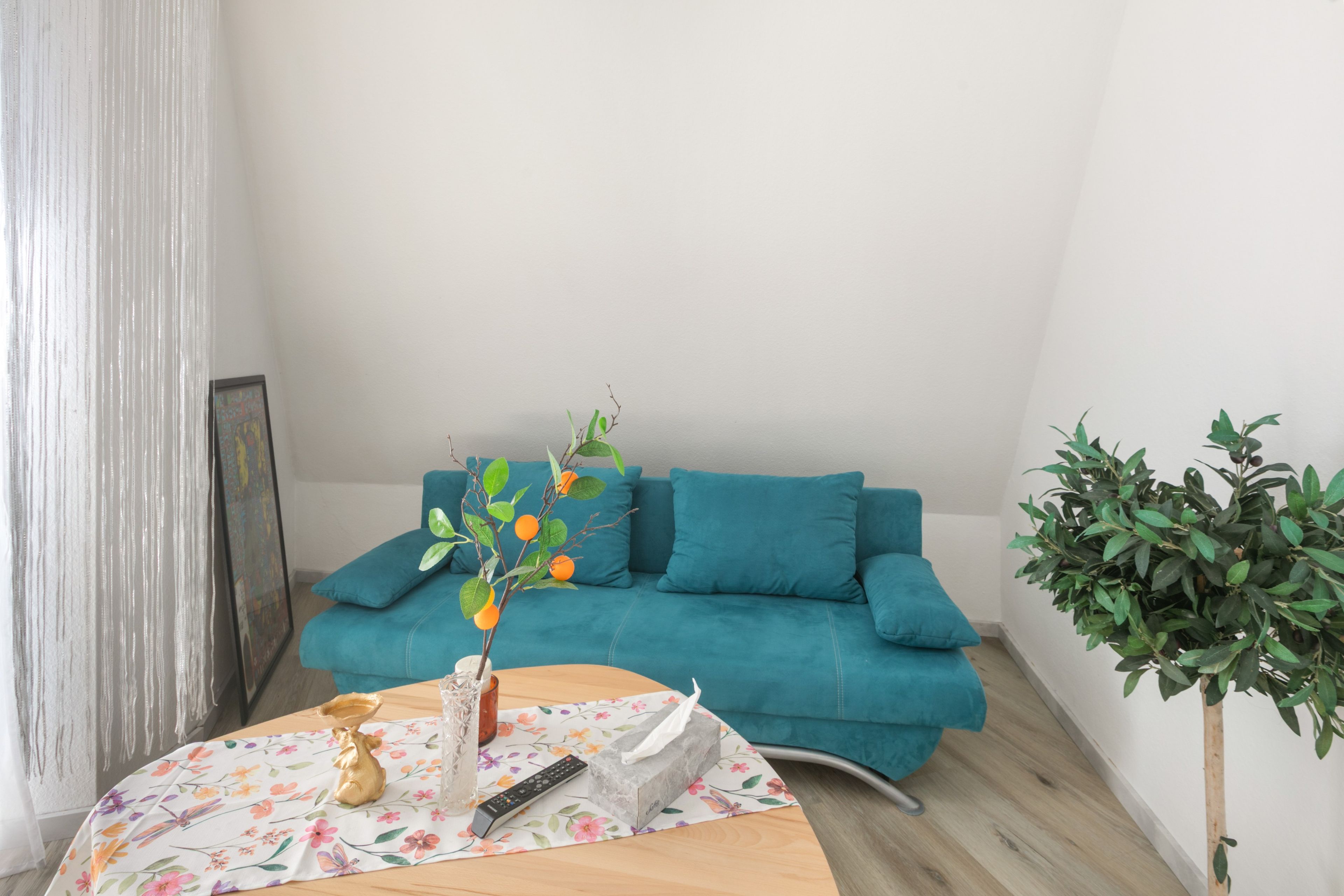 Ein Zimmer mit einem petrolfarbenen Sofa, einem Holztisch mit floralem Tischläufer, einer Pflanze in einer Vase, einer Fernbedienung und einem gerahmten Bild an der Wand.