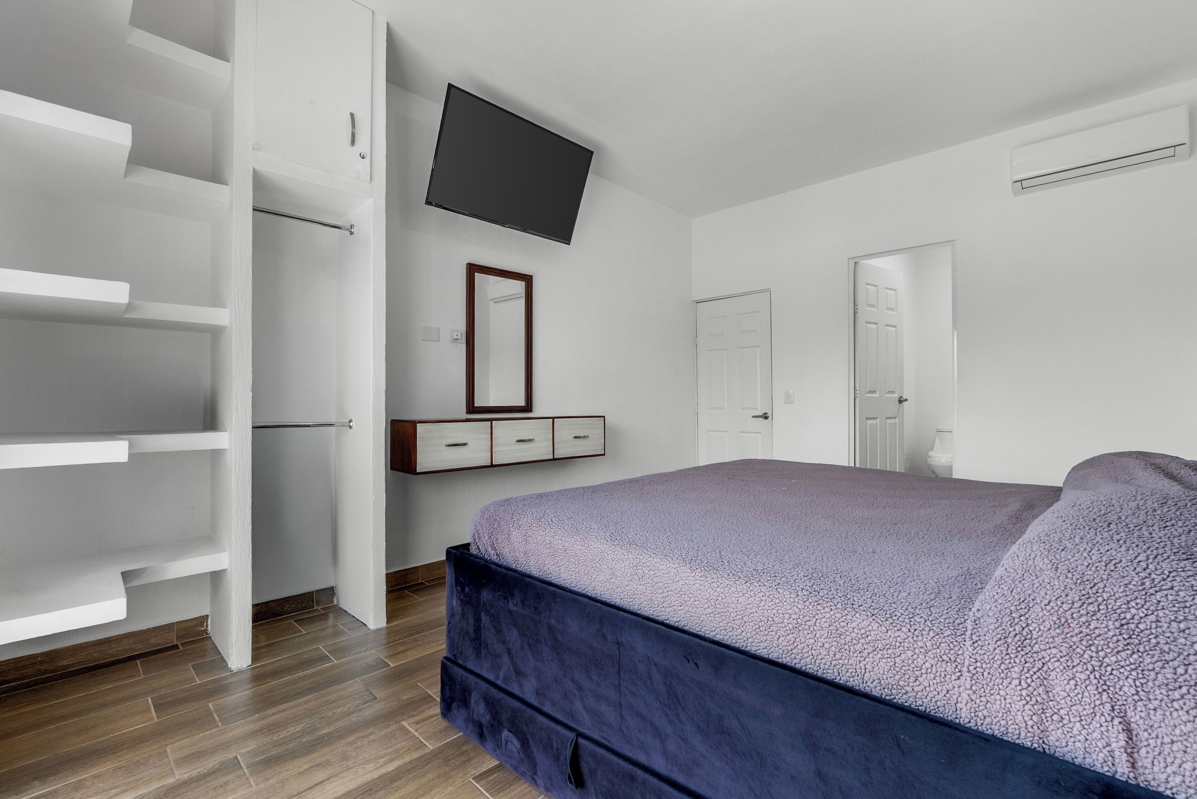 Een slaapkamer met een bed, een kledingkast, een aan de muur bevestigde televisie en een deur die naar een andere kamer leidt.