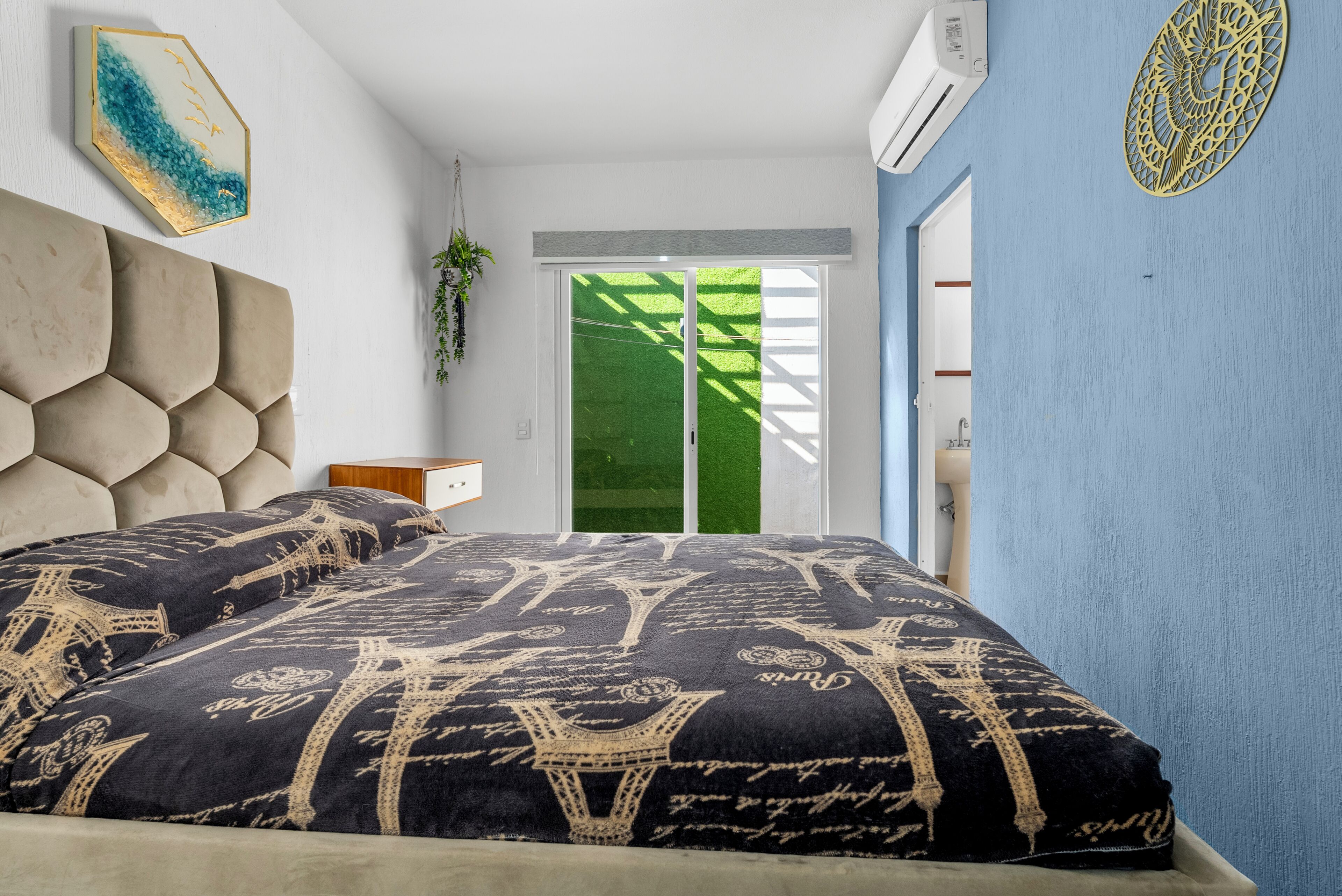 Un dormitorio con una cama grande, aire acondicionado de pared, una ventana con vista a un césped verde y un elemento decorativo en la pared.