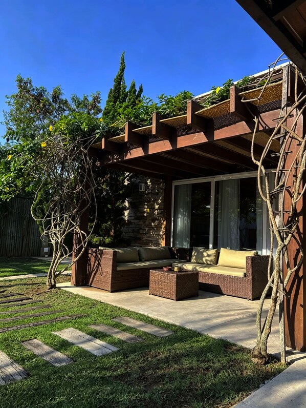 An outdoor patio with a wicker sofa and coffee table, a pergola with climbing plants, and a stone wall backdrop.
