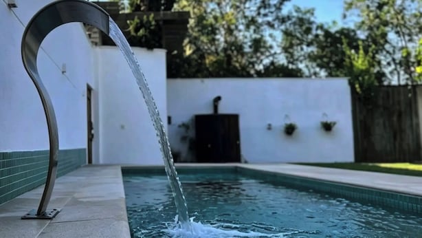 A swimming pool with a curved metal structure at the edge, surrounded by a white wall and greenery.