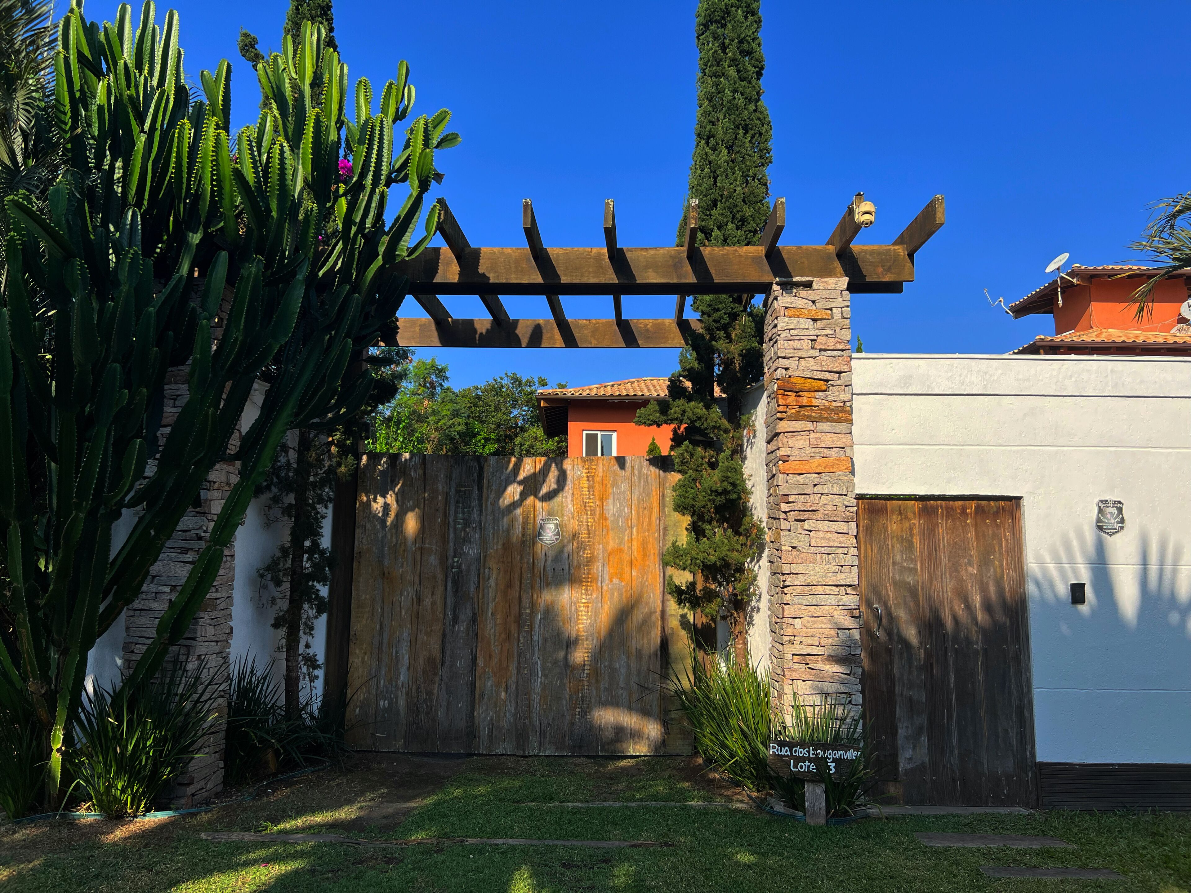 A gated area with a wooden pergola, a stone wall, and a sign indicating 'Rua dos Banguinhos, Lote 3'.