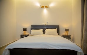 A hotel room with a bed, two bedside tables, and a wall-mounted light fixture. - Cozy 1-bedroom apartment in Abidjan with AC (Abidjan)