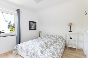 Un dormitorio con cama, ventana con cortinas, un cuadro colgado en la pared, mesita de noche con lámpara y una pequeña estantería.