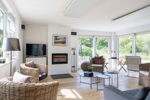 Un salon spacieux, doté de meubles en osier, d’un téléviseur à écran plat et d’une grande fenêtre donnant sur un paysage verdoyant.