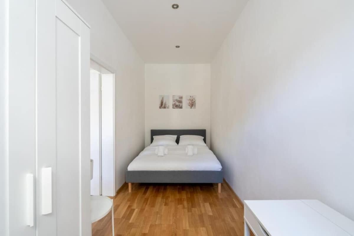 A bedroom with a bed, a white bedside table, and a wooden floor.