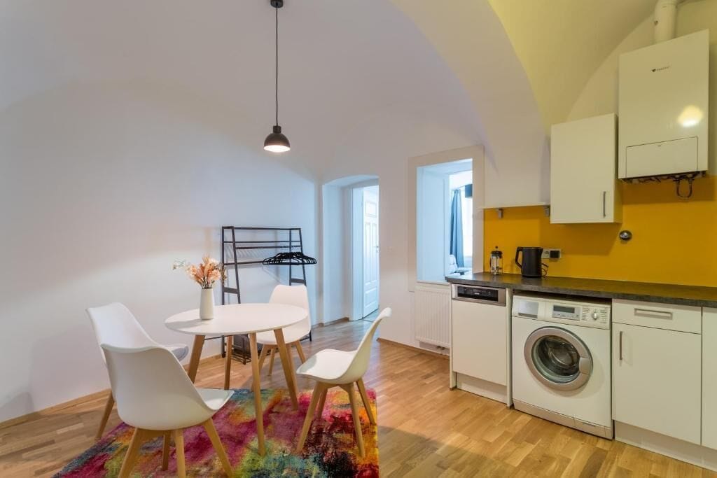 Ett kompakt kök med tvättmaskin, en matplats med bord och stolar samt en hängande torkställning.