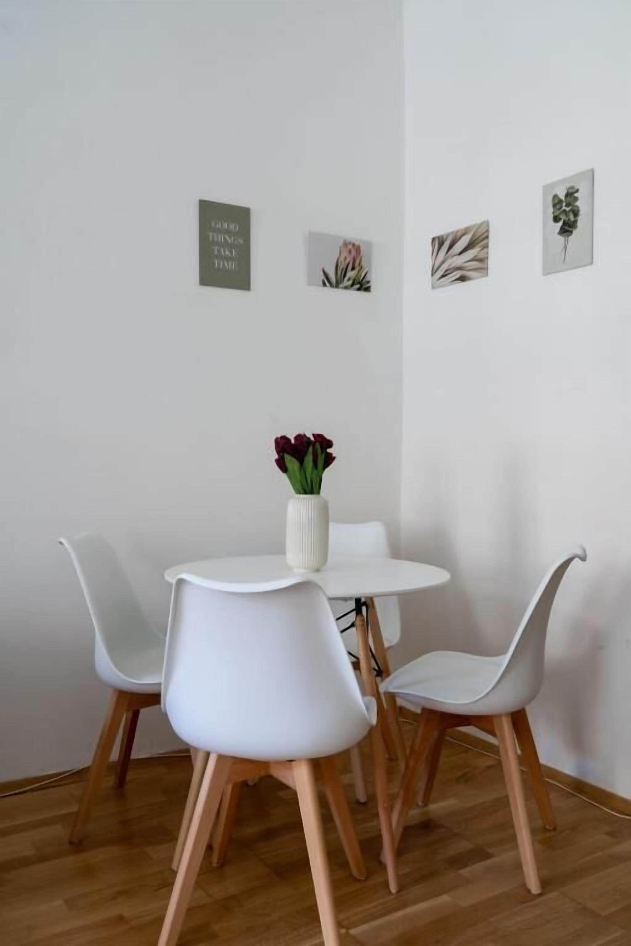 A dining area with a round table, four white chairs, and a vase with red flowers.