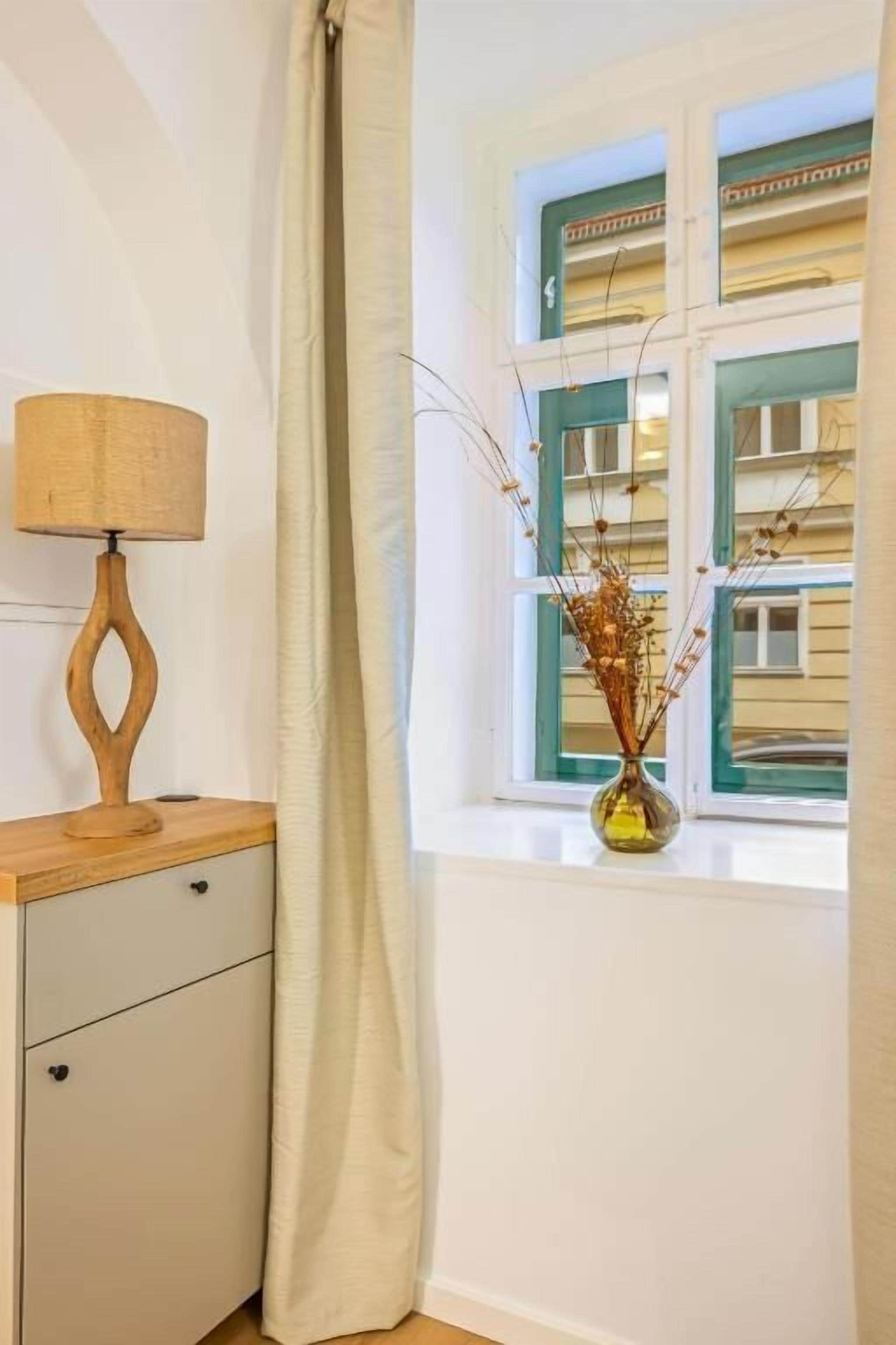 A room with a window, a lamp, a vase with dried plants, and a small wooden cabinet.