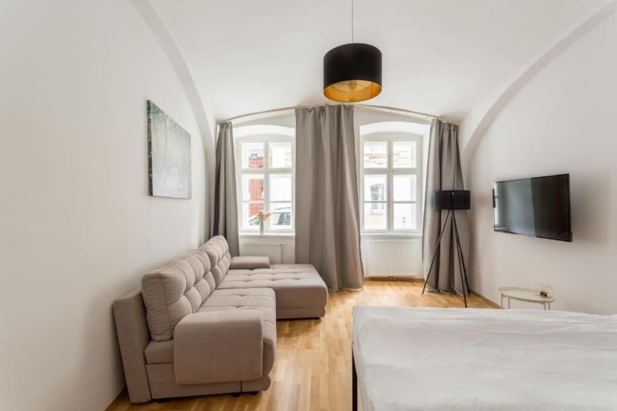 A modern bedroom with a sectional sofa, a bed, a television, and a large window with curtains.