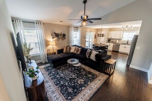 A modern living room with a dark-colored sofa, a coffee table, and a patterned rug. There is a kitchen area visible in the background with white cabinets and stainless steel appliances. - The Grove Getaway - Modern Poolside Retreat (China Grove)