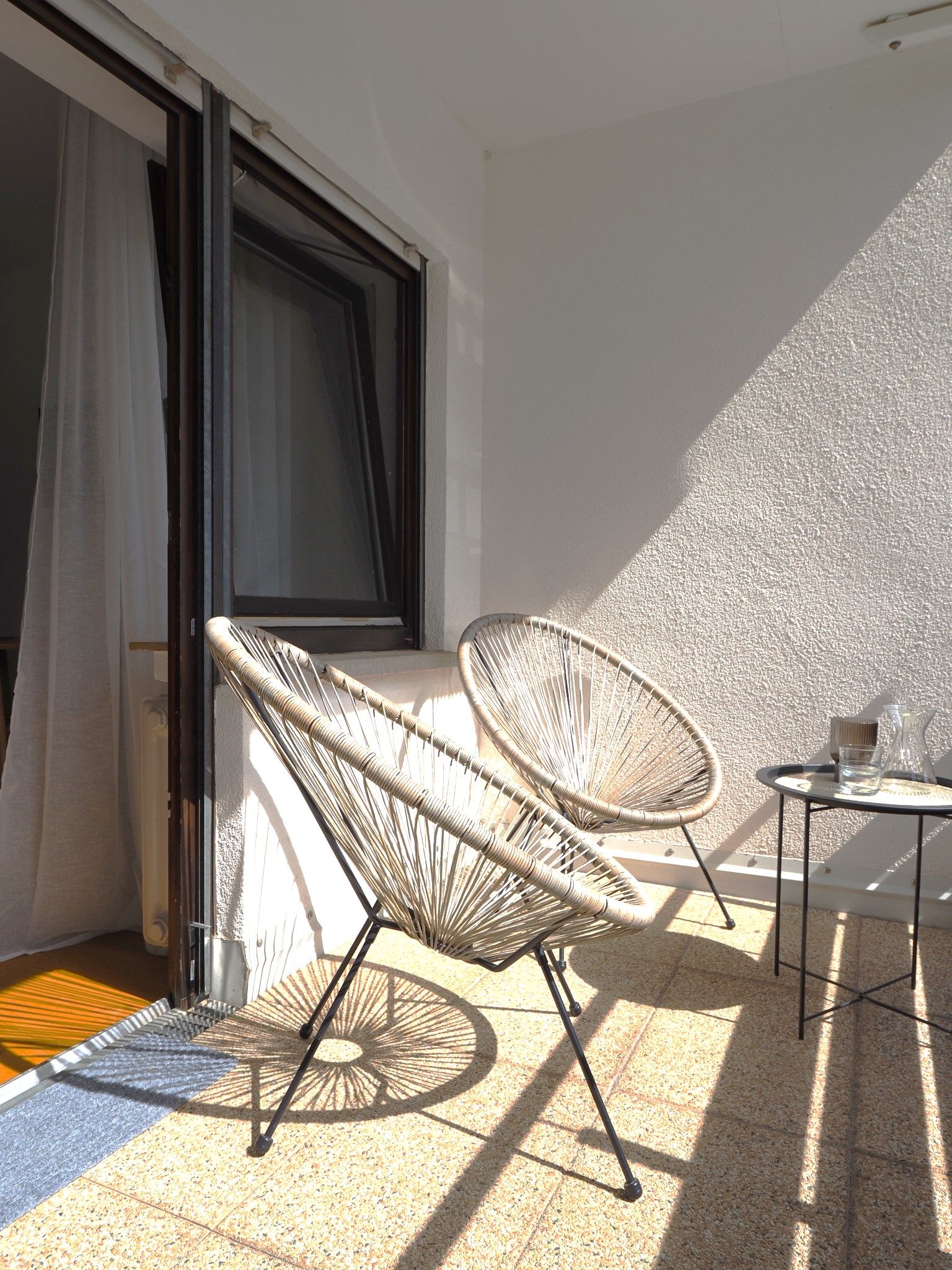 Ein Balkon mit zwei Korbstühlen, einem kleinen Tisch, einem Fenster mit Vorhängen und einer strukturierten Wand.