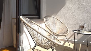 Ein Balkon mit zwei Korbstühlen, einem kleinen Tisch, einem Fenster mit Vorhängen und einer strukturierten Wand.