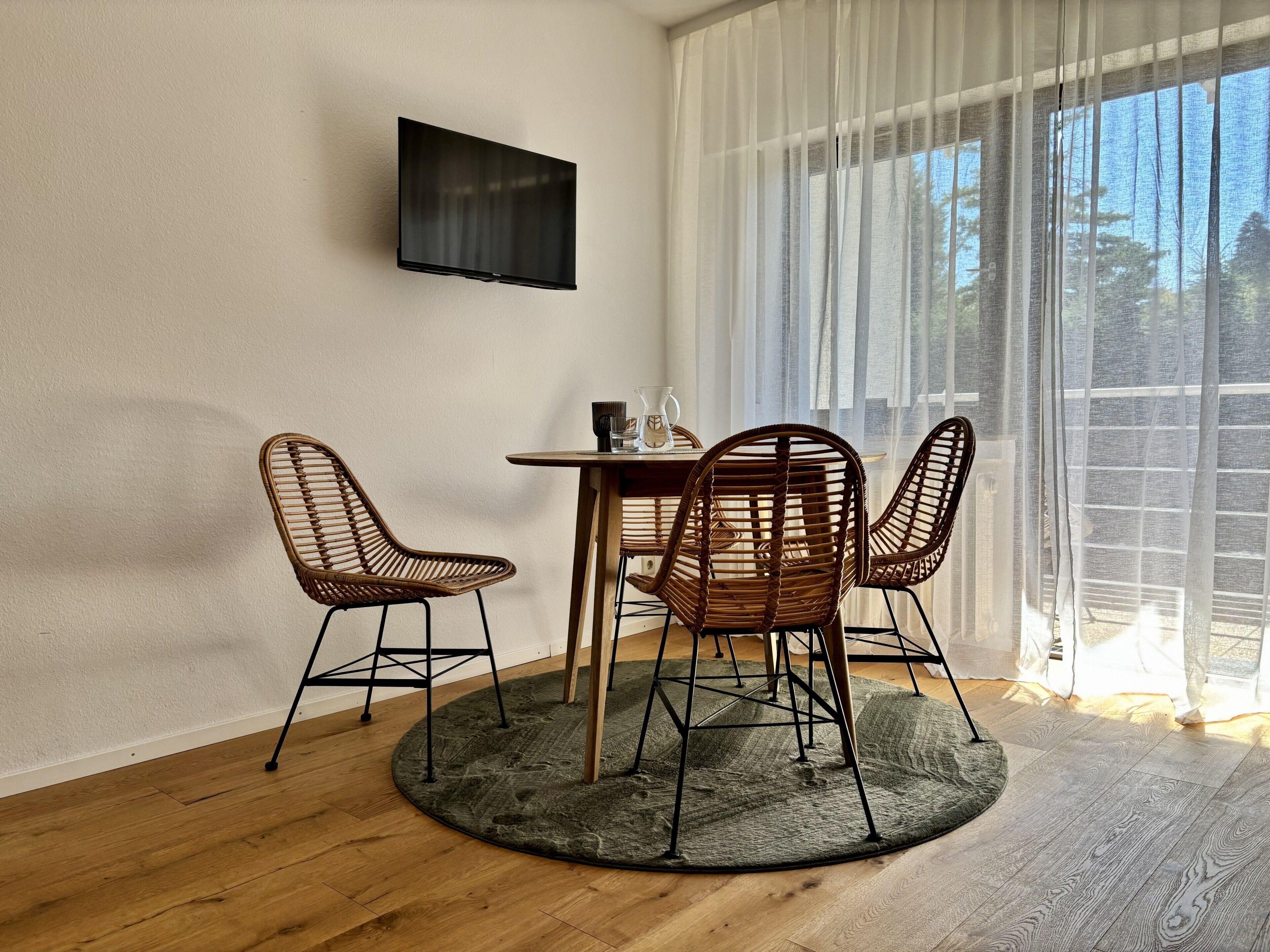 A room with a round table, two wicker chairs, a television on the wall, and a large window with sheer curtains.