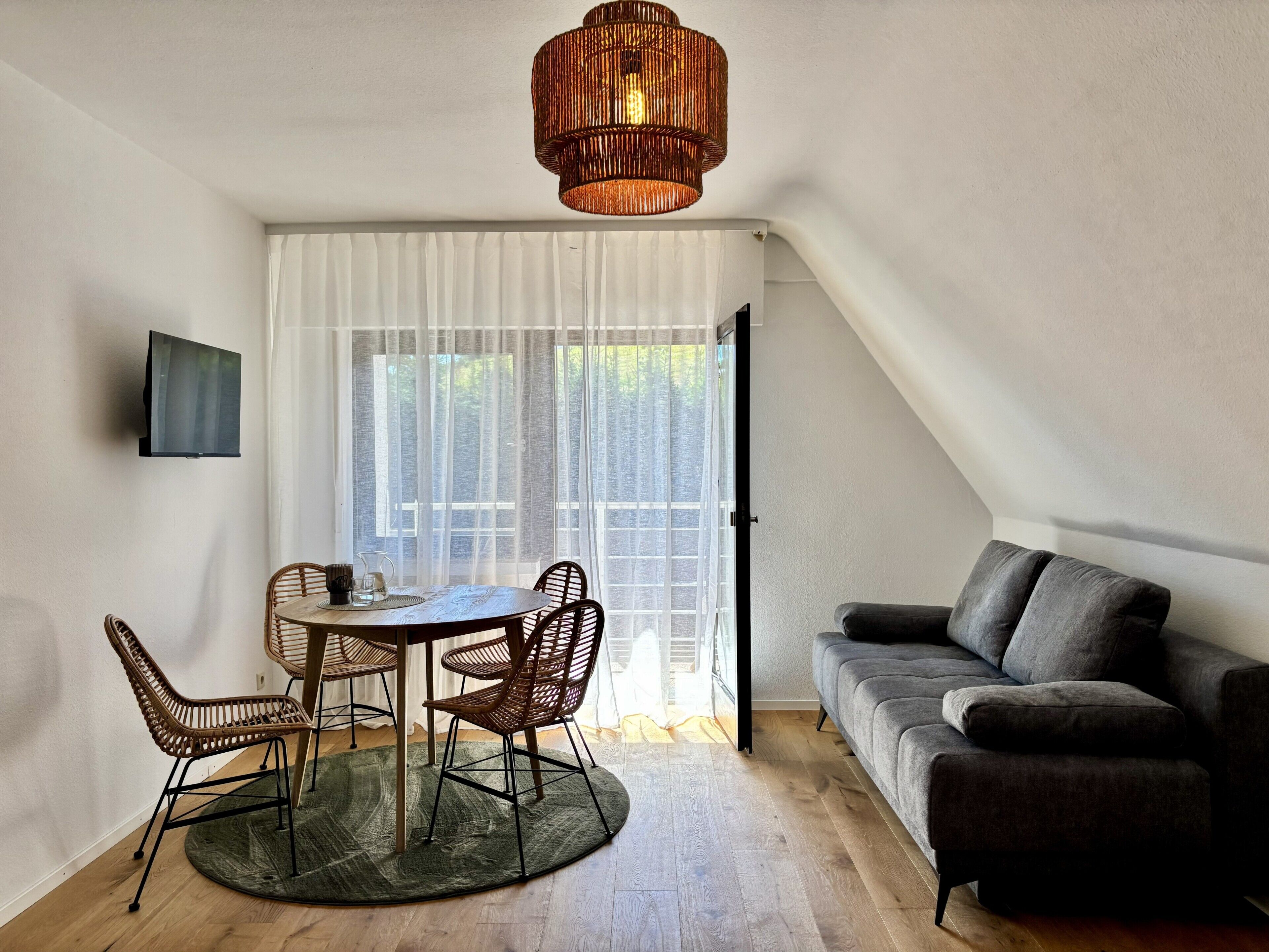 A modern living room with a round table, two chairs, a sofa, a television, and a large window with sheer curtains.