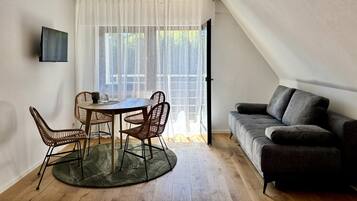 A modern living room with a round table, two chairs, a sofa, a television, and a large window with sheer curtains.