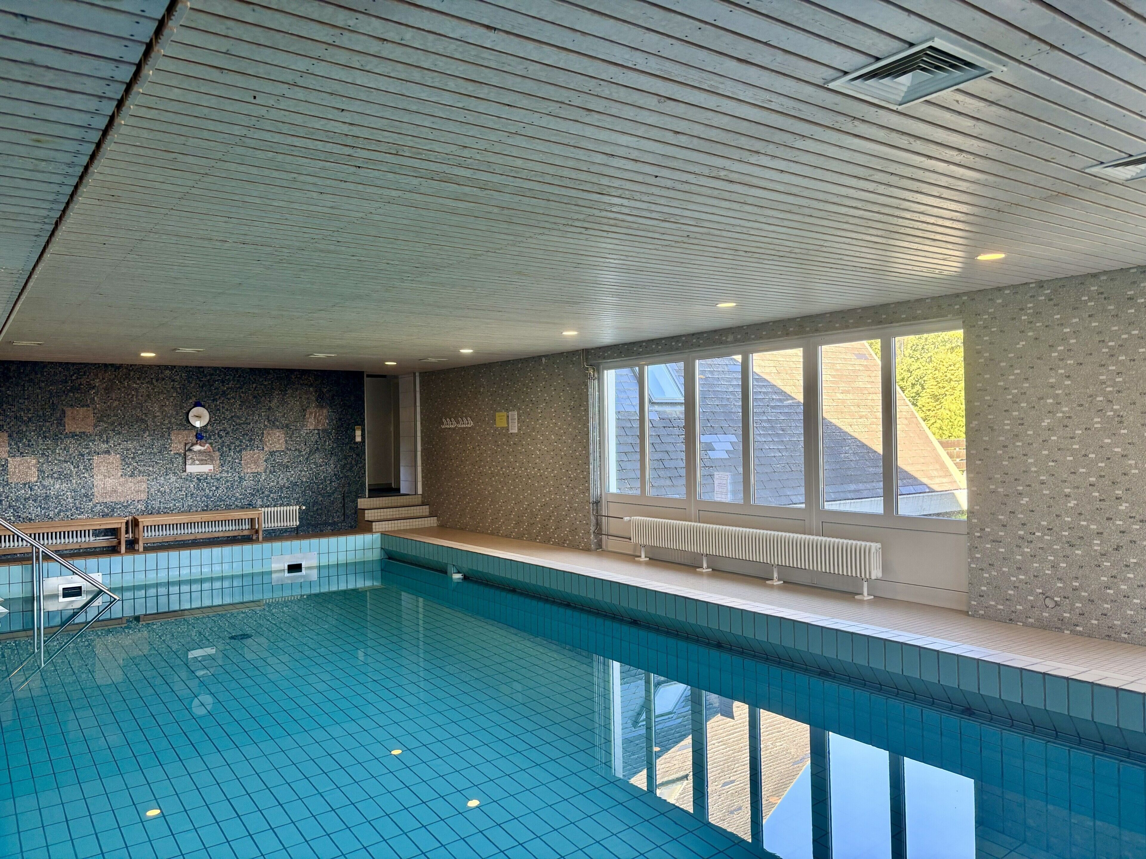 Una piscina al coperto con pareti e soffitto piastrellati, panche lungo il bordo e ampie finestre che lasciano entrare luce naturale.