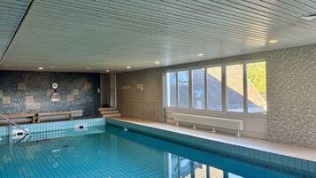 Una piscina al coperto con pareti e soffitto piastrellati, panche lungo il bordo e ampie finestre che lasciano entrare luce naturale.